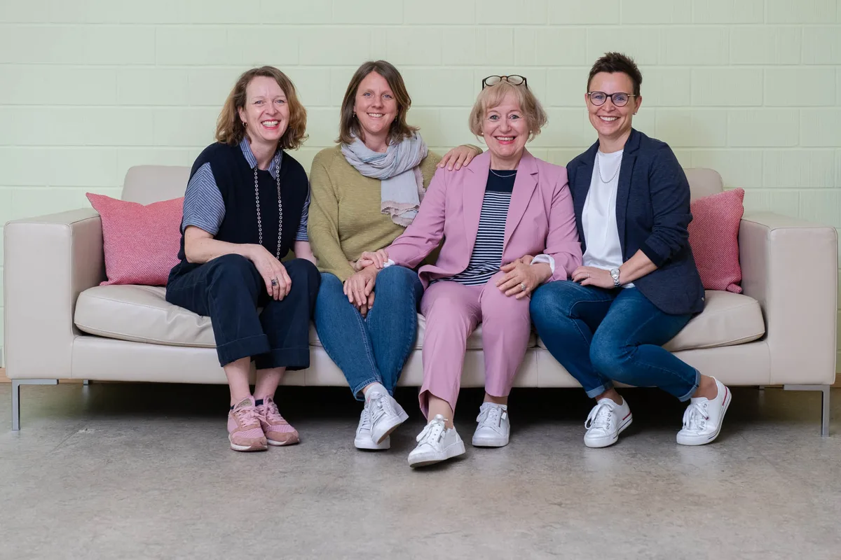Vier Frauen sitzen vertraut auf einem Sofa und lächeln in die Kamera.